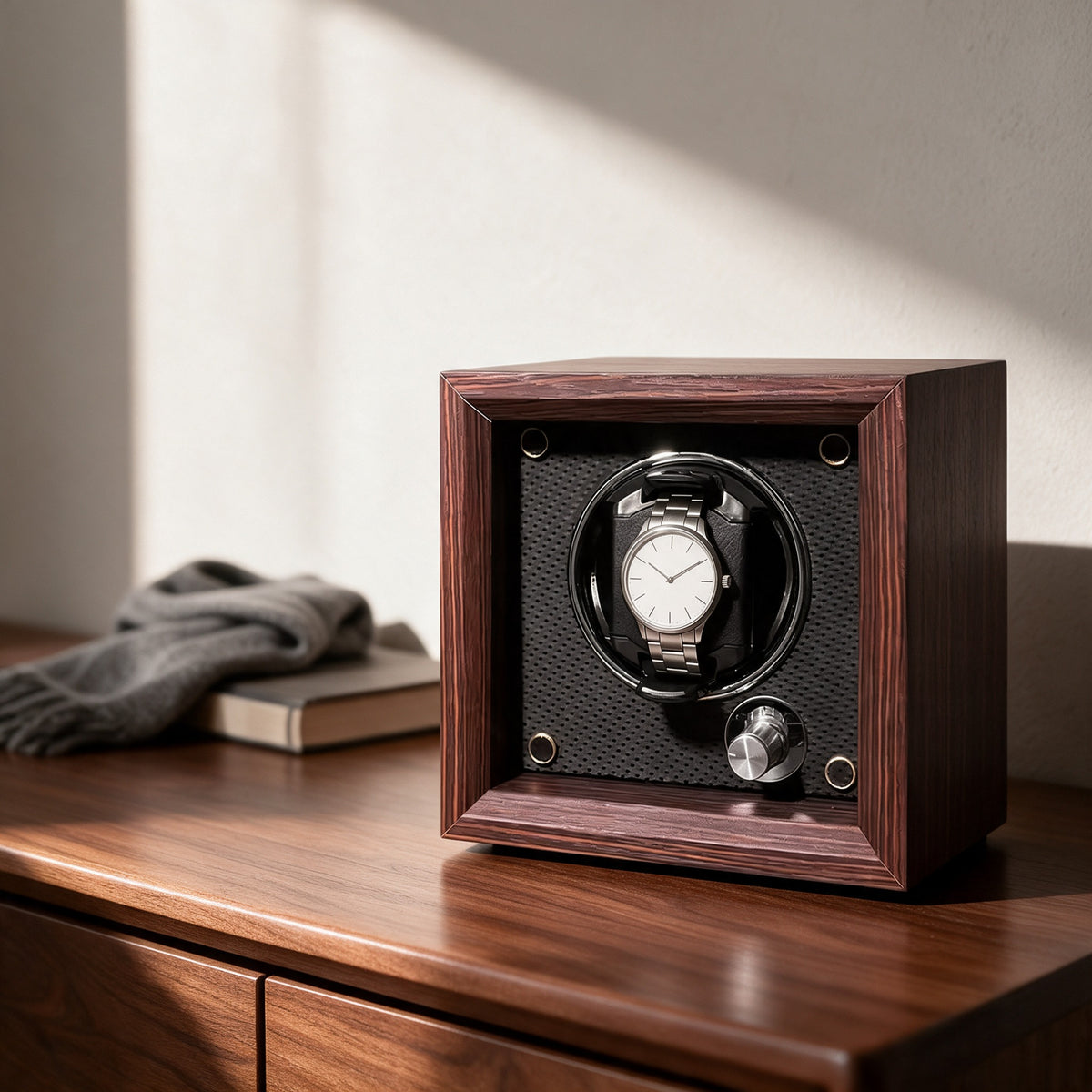 The Whitmore watch winder on the walnut top of a chest of drawers, beside a folded wool scarf and a book.