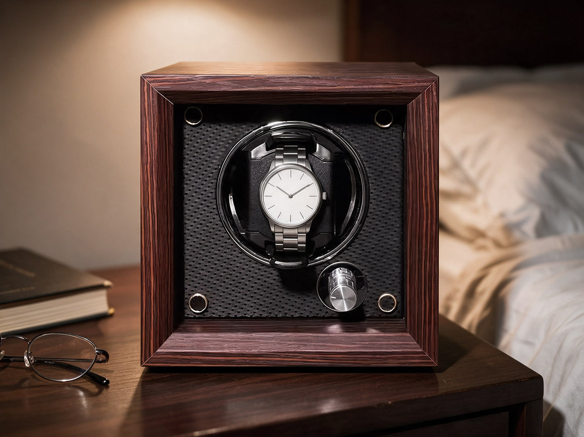 The Whitmore watch winder in black walnut veneer on a bedside table at night, a warm lamp to one side, reading glasses and a book beside it, the edge of a bed behind.